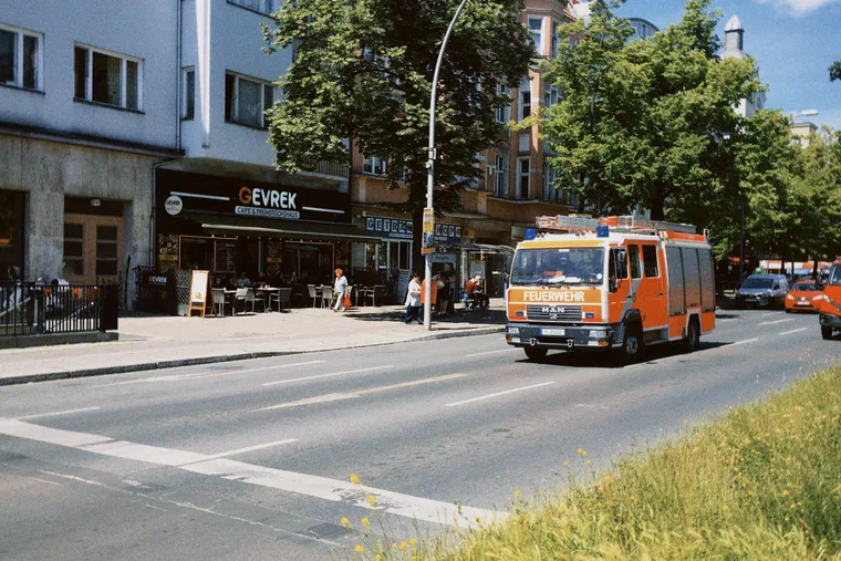 A Berlin street in summer, with a café, parked cars and a passing fire engine.
