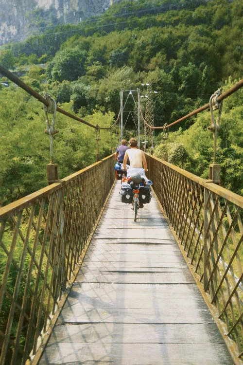 Two cyclists crossing a narrow old wooden suspension footbridge over a wooded gorge.