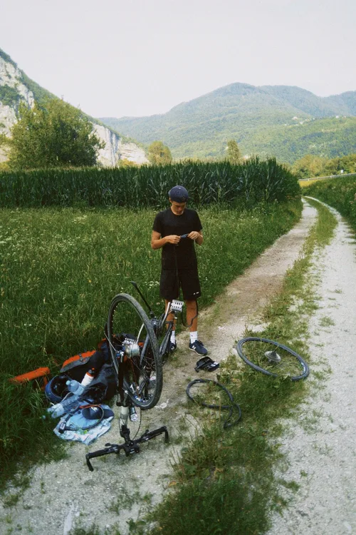A cyclist fixing a puncture beside a gravel path, his bike upended and a wheel laid out on the ground, with a cornfield and mountains behind.