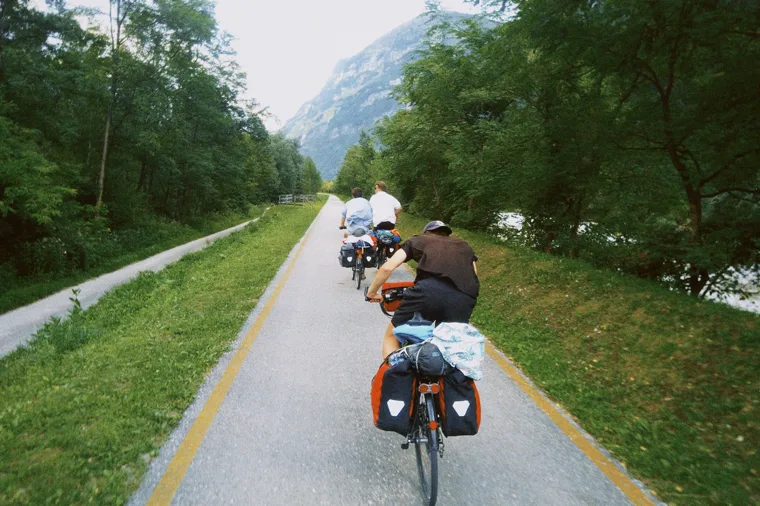 Three cyclists riding single file along a forest cycle path towards a steep mountain.